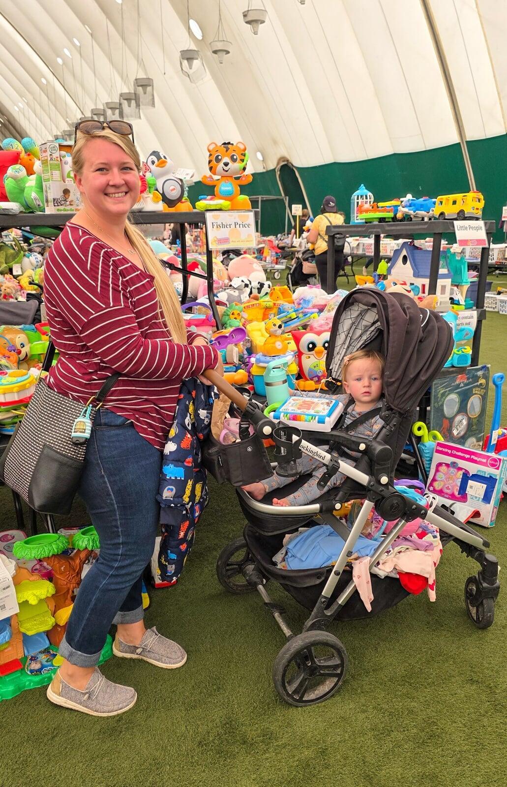 A mom with a large JBF shopping bag on her shoulder stands beside her husband who wears their toddler at a JBF sale.